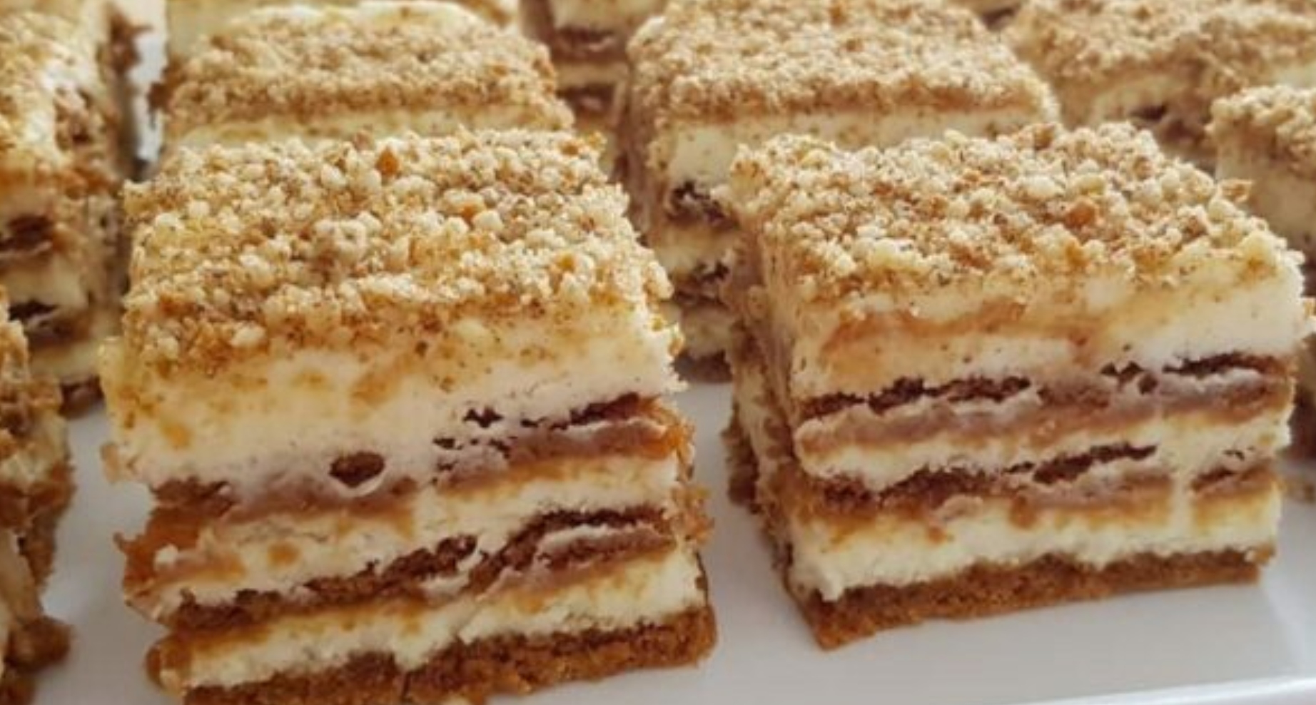 Vrstvené nepečené řezy ze sušenek Lotus Biscoff s krémem z mascarpone, posypané drcenými sušenkami a ořechy na servírovacím talíři.