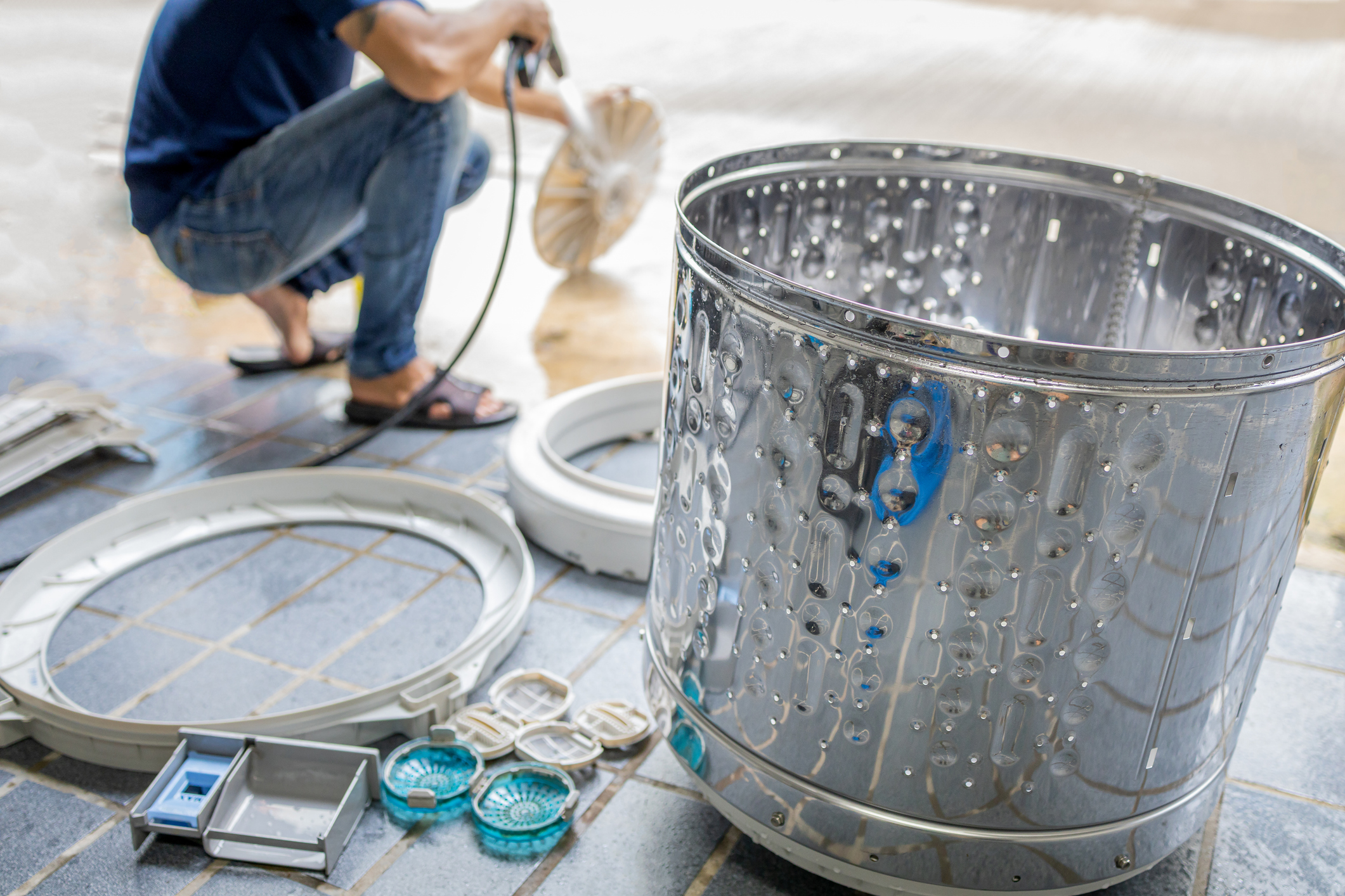 Rozložená pračka s vyndaným bubnem a díly na zemi pro čištění.