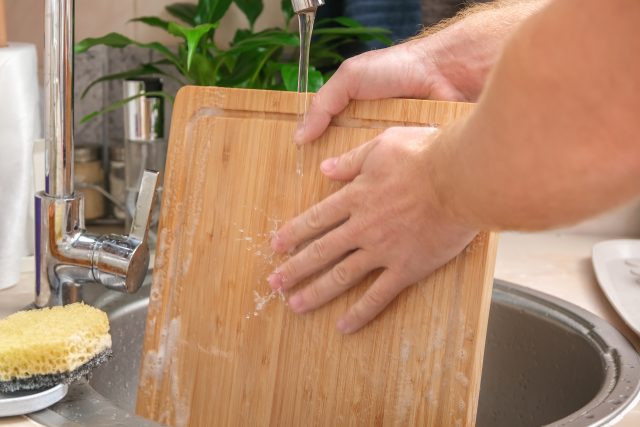 A man washes wooden bamboo cutting board in the kitchen sink under running water. Gentle hand washing of a wooden cutting board. Cleaning dirty kitchen wood products.