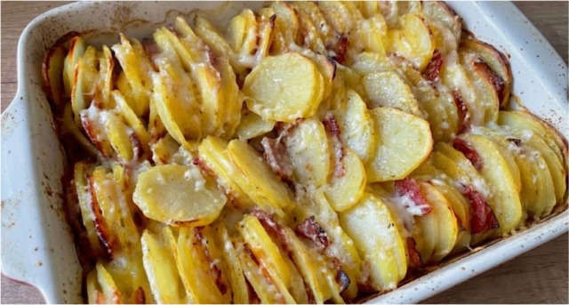 Detailní pohled na zapékací mísu s vertikálně naskládanými plátky brambor a slaniny, se zlatavým rozpuštěným sýrem a křupavými okraji.
