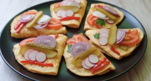 Smažené vaječné omelety na černém talíři obložené čerstvou zeleninou, ředkvičkami, cibulí a kyselou okurkou