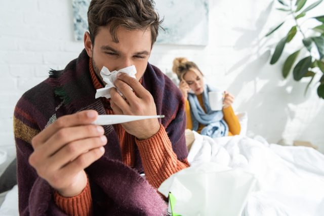 sick man with fever holding thermometer and napkin in bedroom with woman behind