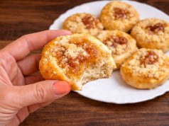 Detail nakousnutého domácího koláčku s tvarohem, marmeládou a drobenkou drženého v ruce, v pozadí talíř s upečenými koláčky.