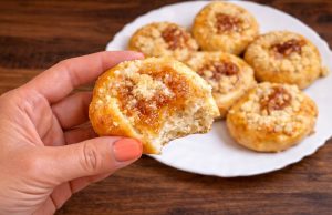 Detail nakousnutého domácího koláčku s tvarohem, marmeládou a drobenkou drženého v ruce, v pozadí talíř s upečenými koláčky.