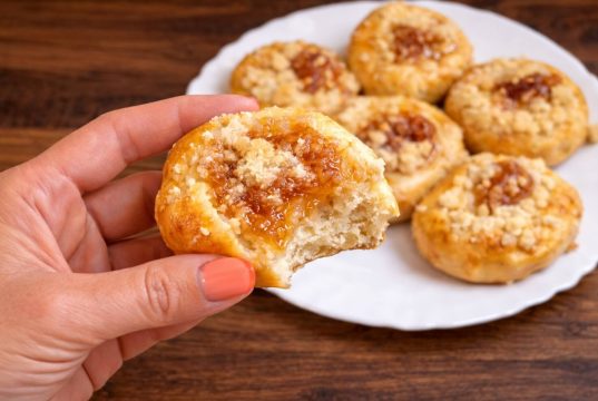 Detail nakousnutého domácího koláčku s tvarohem, marmeládou a drobenkou drženého v ruce, v pozadí talíř s upečenými koláčky.