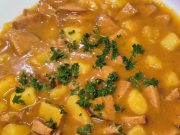 Nejlepší domácí buřtguláš: Hustá omáčka a dokonalá chuť díky základu na sádle Detailní pohled na talíř plný poctivého českého buřtguláše. V husté červenohnědé omáčce jsou vidět kousky brambor a nakrájených opékaných buřtů, celá porce je posypaná čerstvou nasekanou petrželkou.