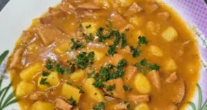 Nejlepší domácí buřtguláš: Hustá omáčka a dokonalá chuť díky základu na sádle Detailní pohled na talíř plný poctivého českého buřtguláše. V husté červenohnědé omáčce jsou vidět kousky brambor a nakrájených opékaných buřtů, celá porce je posypaná čerstvou nasekanou petrželkou.