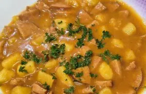 Nejlepší domácí buřtguláš: Hustá omáčka a dokonalá chuť díky základu na sádle Detailní pohled na talíř plný poctivého českého buřtguláše. V husté červenohnědé omáčce jsou vidět kousky brambor a nakrájených opékaných buřtů, celá porce je posypaná čerstvou nasekanou petrželkou.