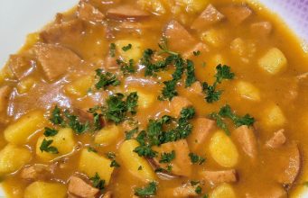 Nejlepší domácí buřtguláš: Hustá omáčka a dokonalá chuť díky základu na sádle Detailní pohled na talíř plný poctivého českého buřtguláše. V husté červenohnědé omáčce jsou vidět kousky brambor a nakrájených opékaných buřtů, celá porce je posypaná čerstvou nasekanou petrželkou.