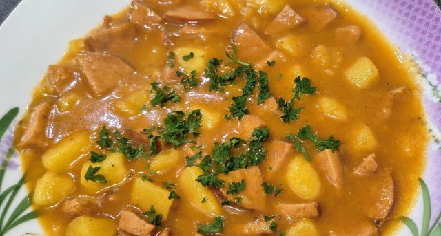 Detailní pohled na talíř plný poctivého českého buřtguláše. V husté červenohnědé omáčce jsou vidět kousky brambor a nakrájených opékaných buřtů, celá porce je posypaná čerstvou nasekanou petrželkou.