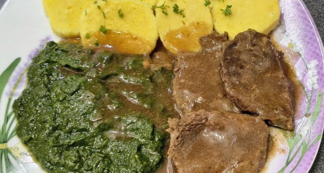 Plátky pečeného hovězího masa přelité tmavým sosem na talíři, doplněné o porci zeleného krémového špenátu a plátky bramborového knedlíku.