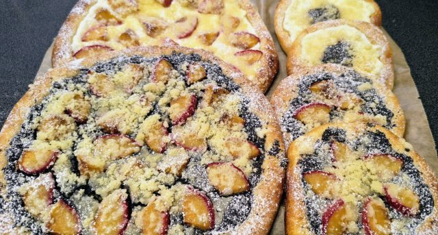Dva velké a několik menších čerstvě upečených kynutých koláčů na pečicím papíře. Zdobené jsou makovou a tvarohovou náplní, plátky slív a bohatě posypané máslovou drobenkou a moučkovým cukrem.