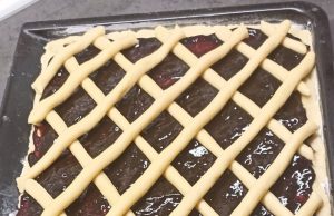 Tradiční mřížkový koláč s povidly: Bleskové křehké těsto z robotu Čerstvě upečený křehký koláč na plechu s bohatou vrstvou tmavých švestkových povidel, který je na povrchu ozdobený pravidelnou mřížkou ze zlatavě upečeného těsta.