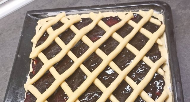 Čerstvě upečený křehký koláč na plechu s bohatou vrstvou tmavých švestkových povidel, který je na povrchu ozdobený pravidelnou mřížkou ze zlatavě upečeného těsta.