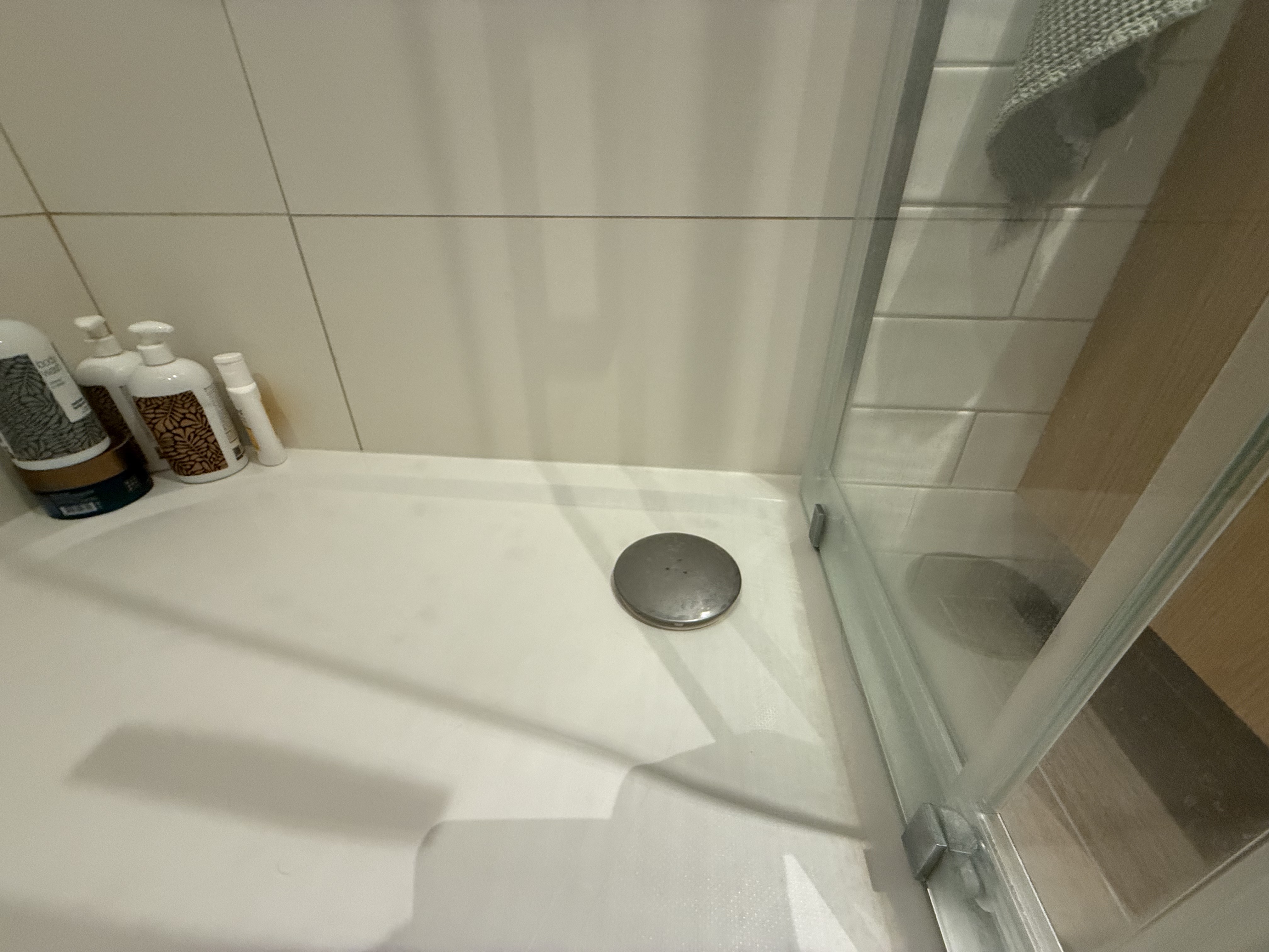 Bílý sprchový kout s pootevřenými skleněnými dveřmi a detailem odtokové mřížky, vedle stojí lahvičky s kosmetikou.