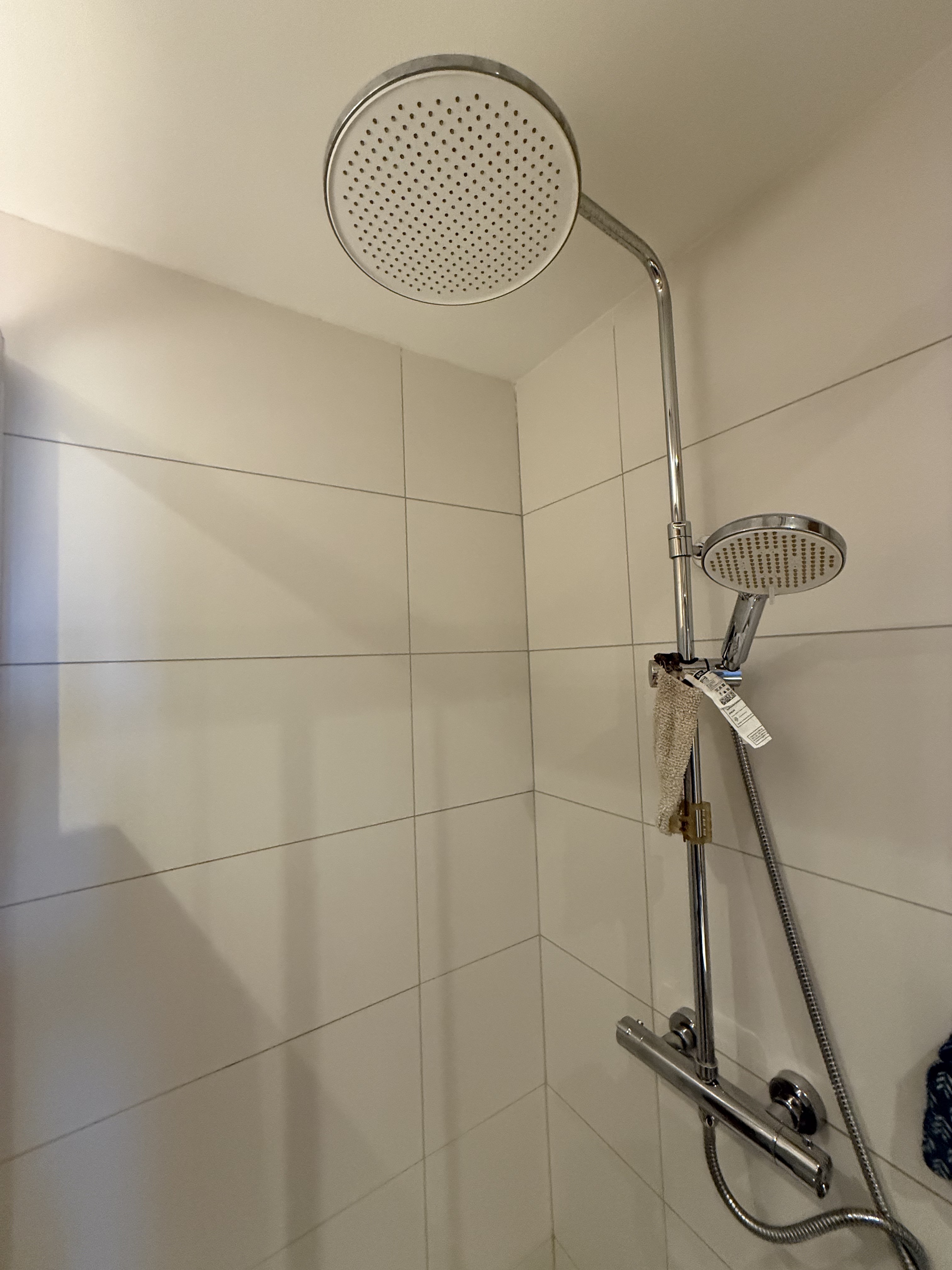Detail moderní chromované sprchové soupravy s hlavovou a ruční sprchou na bílé kachličkové stěně.
