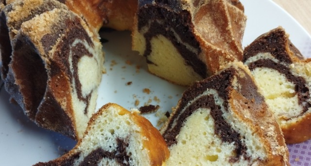 Domácí mramorová bábovka s tvarohem na bílém talíři, nakrájená na plátky, s viditelnou jemnou strukturou a kakaovým vzorem na dřevěném stole.