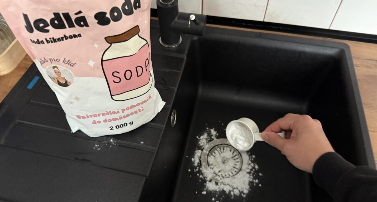 Sypání jedlé sody pomocí odměrky do odtoku černého granitového dřezu.