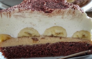 Luxusní banánový dort s arašídovým máslem a mascarpone: Chuťová exploze v každém soustu Detailní řez banánovým dortem s viditelnými vrstvami tmavého arašídového krému, celými banány a vysokou čepicí bílého mascarpone krému.