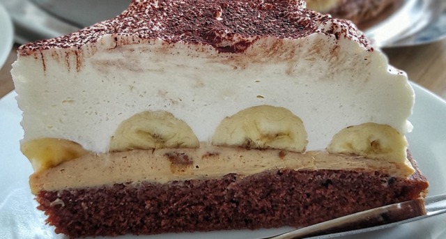 banánový dezert (2) Detailní řez banánovým dortem s viditelnými vrstvami tmavého arašídového krému, celými banány a vysokou čepicí bílého mascarpone krému.
