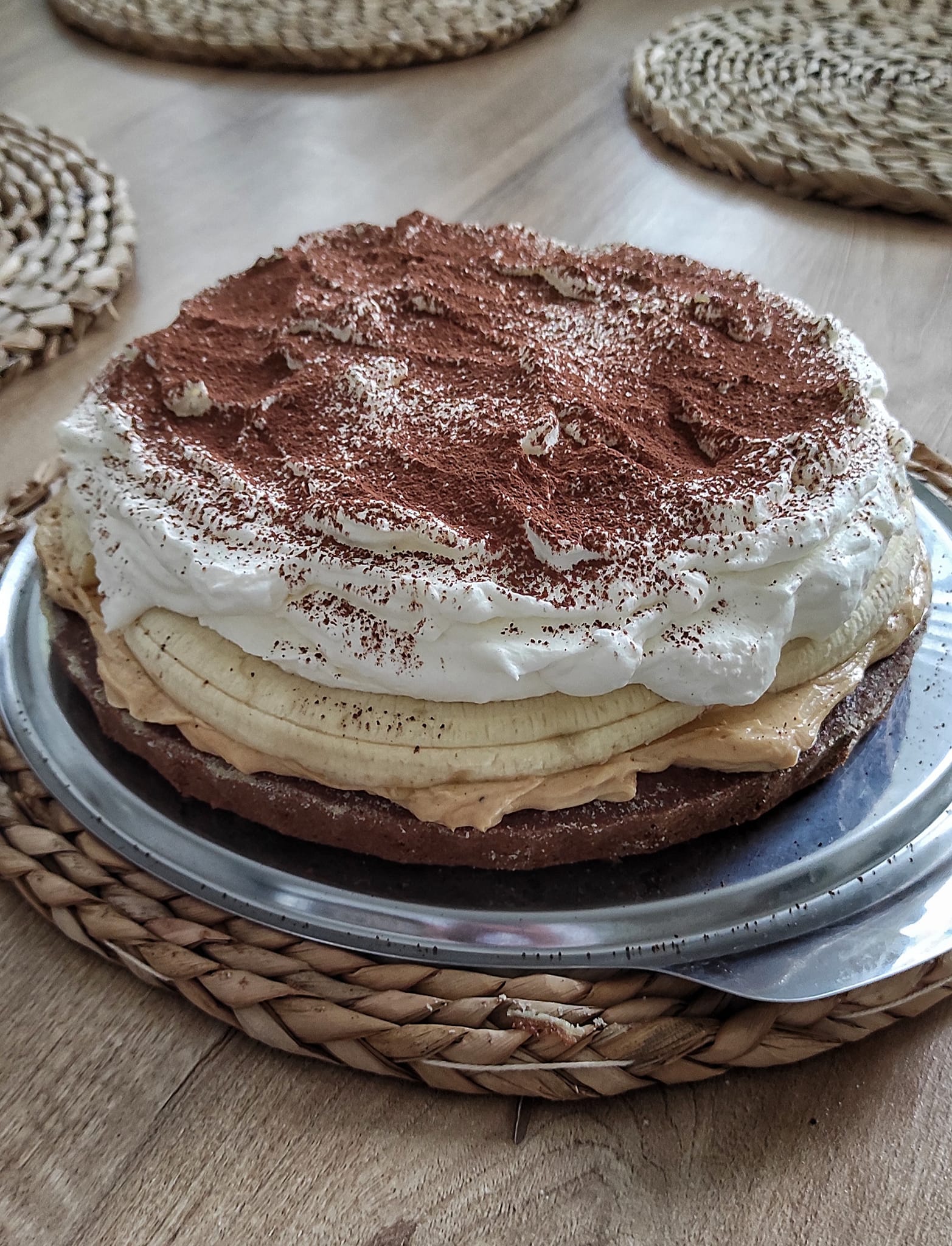 Detailní řez banánovým dortem s viditelnými vrstvami tmavého arašídového krému, celými banány a vysokou čepicí bílého mascarpone krému.