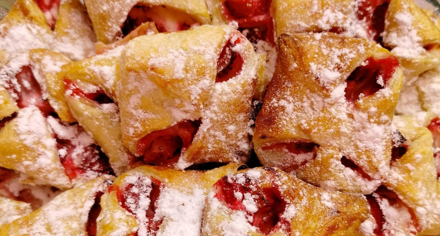 Detailní záběr na malé čtvercové taštičky z listového těsta plněné jahodami, štědře posypané moučkovým cukrem.